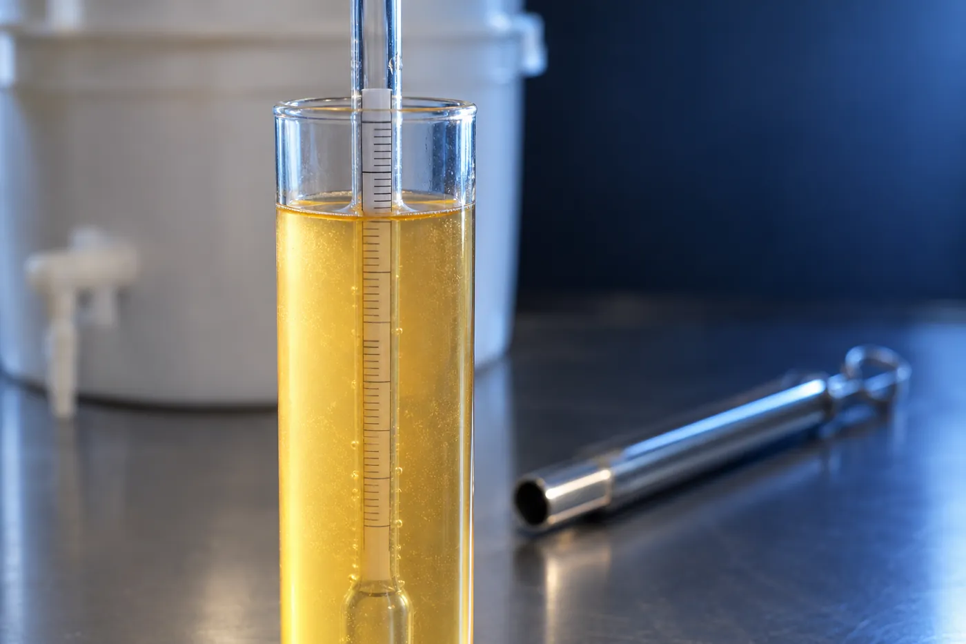 A glass hydrometer floating in a tall trial jar of golden, slightly cloudy wash, seen at the level of the surface.